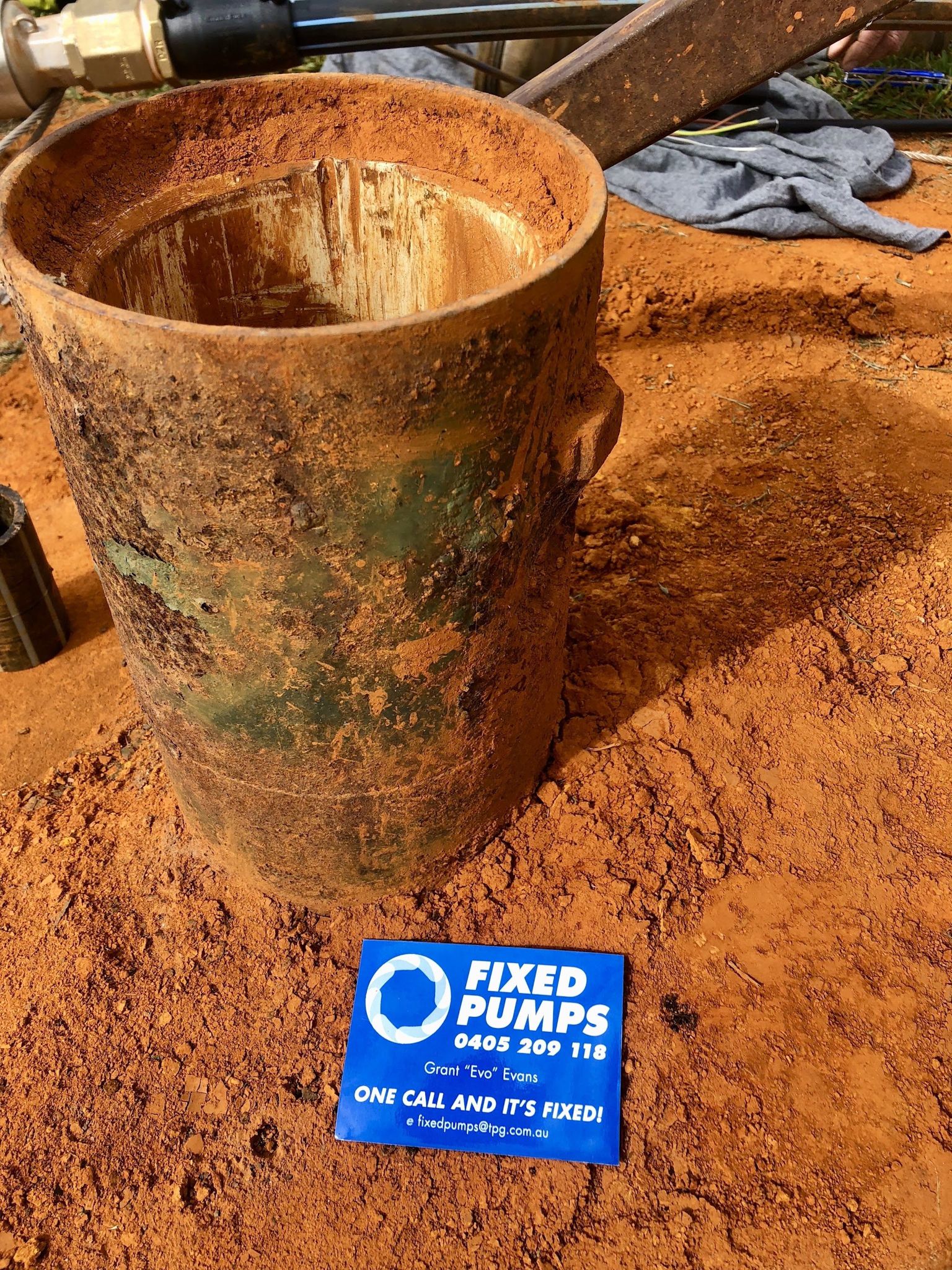 A rusted bore casing on red soil beside a Fixed Pumps business card