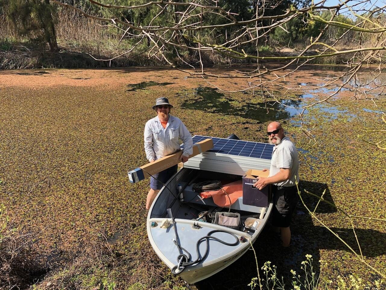 Two men in a tinny on a weedy dam with a solar panel and pump gear